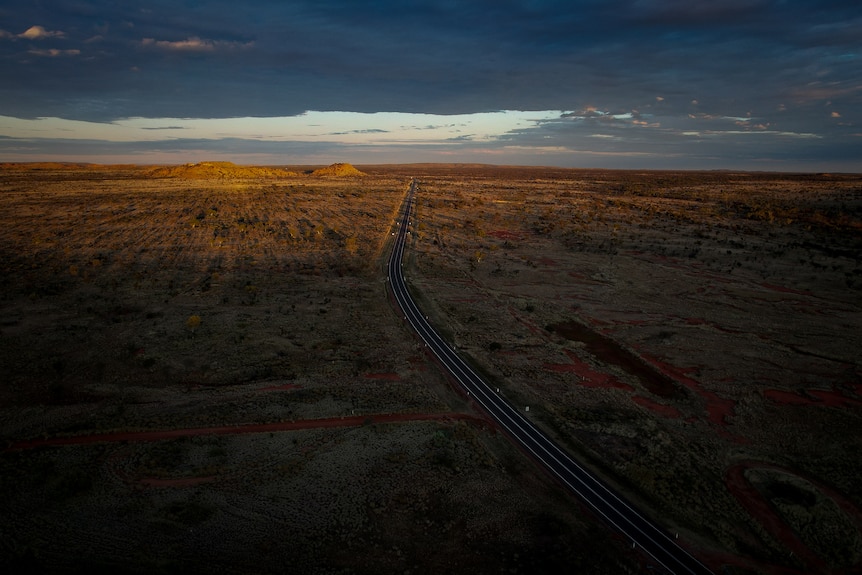 Aerial shot over vast outback landscape shows a dark bitumen highway running through the orange dirt, dotted with green trees.