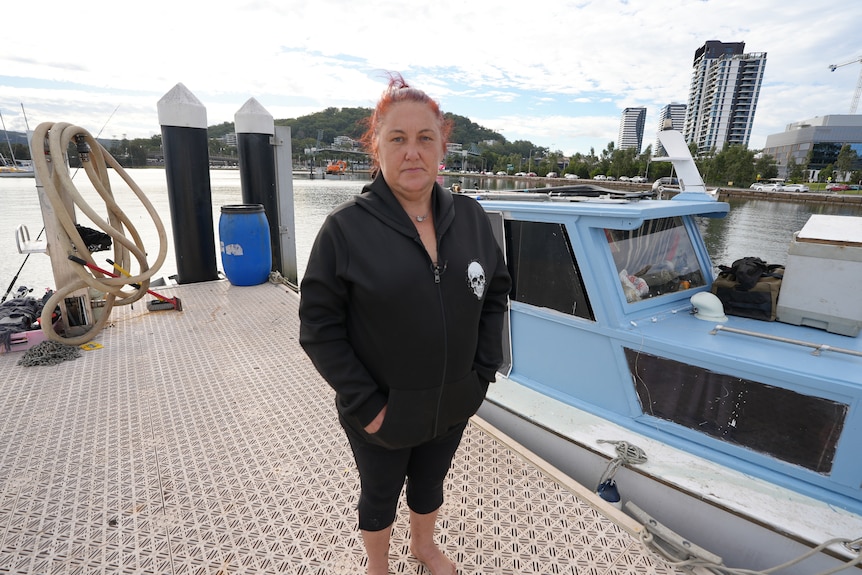 A woman wearing a black hoodie and black pants standing on a wharf next to a blue boat.