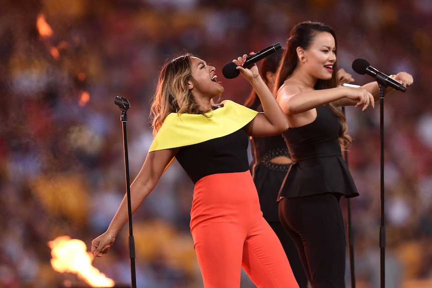 Jess Mauboy on stage singing, leaning back dramatically, wearing a yellow and black top and red pants