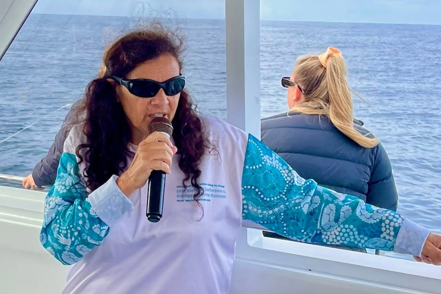 Jodi wears a blue and white long sleeve top and talks into a microphone on a boat.
