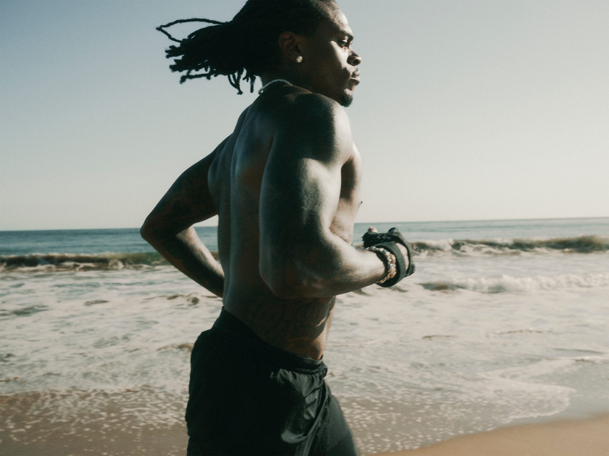 A man with tattoos runs along the shore, with ocean waves crashing in the background under a clear sky.