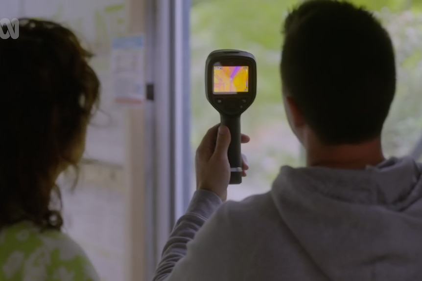 A man holds a thermal camera up to a window to see where air might be coming in from