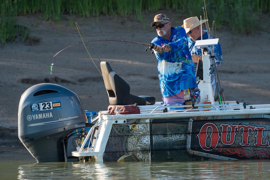 Two white men on a gray small fishing boat, wearing a blue long-sleeve shirt, caps, black glasses, fishing poles over edge