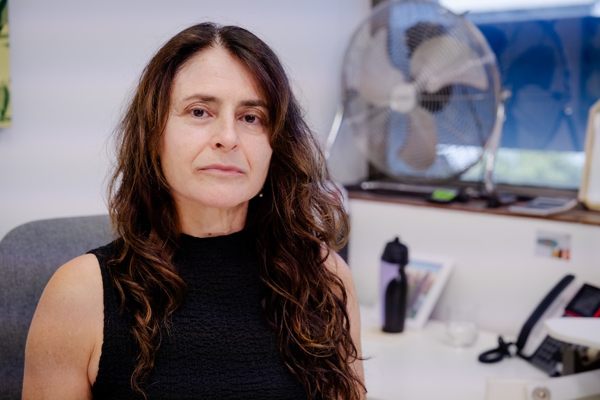 A white woman with long brown hair, wearing a turtle-neck black top, sitting at her desk with a serious expression.