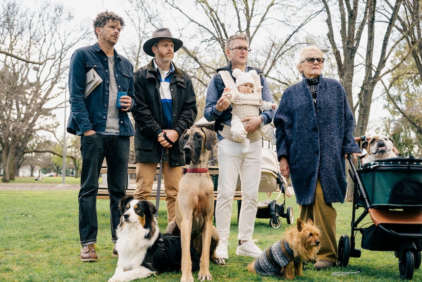 A group of people stand behind dogs in a dog park while watching something off screen.