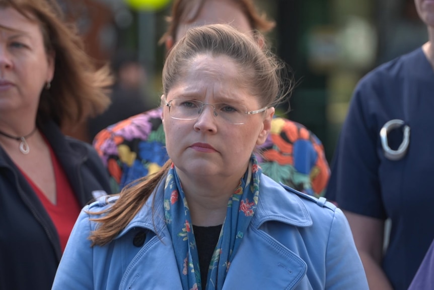 Elizabeth Dabars has brown hair and wears a blue coat, she looks off camera, behind her are women wearing scrubs