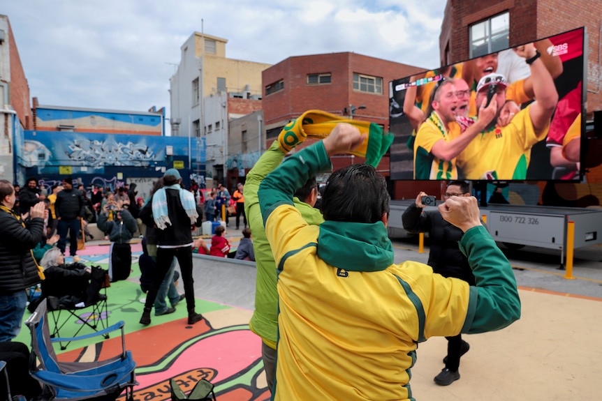 Seen from behind, men wearing green and gold shirts and one holding a scarf, raise their arms as they look at a big screen in f 