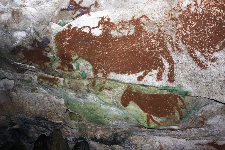 Reddish-brown drawings of human figures and four legged animals adorn a green and grey rocky background.