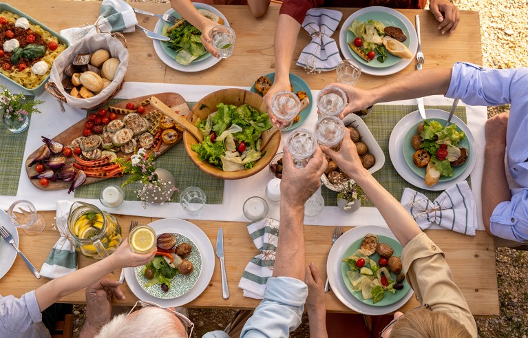Overhead view of a wooden table outdoors, with plates of salad, grilled vegetables and bread. Hands holding drinking glasses are raised in a toast.
