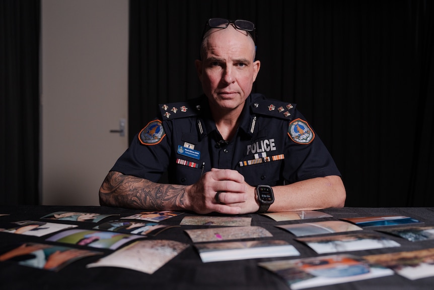 A uniformed police officer sits at a table where photos are laid out, he looks at the camera with a serious expression.