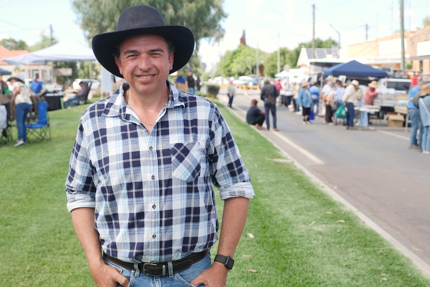 A smiling man in a dark Akubra and checked shirt stands on some grass next to a street in a country town.