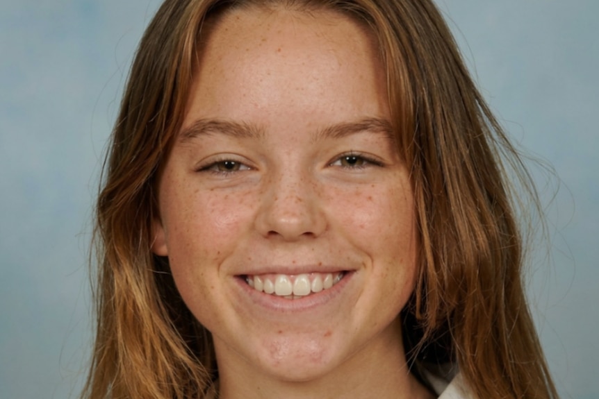 Alcock smiling with caramel coloured long hair and freckles across her cheeks and nose wearing a white school shirt.