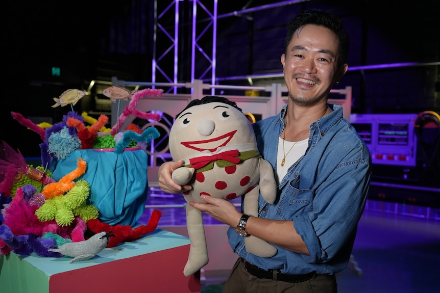 Benjamin Law smiles big holding the Play School toy Humpty. The background is the colourful set