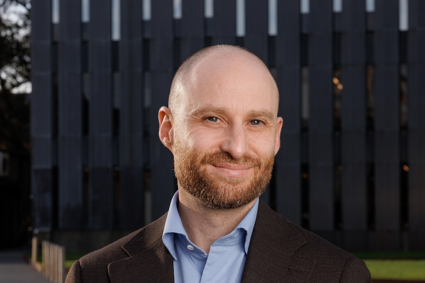 Man in a shirt and brown coat stands outside and smiles at camera.