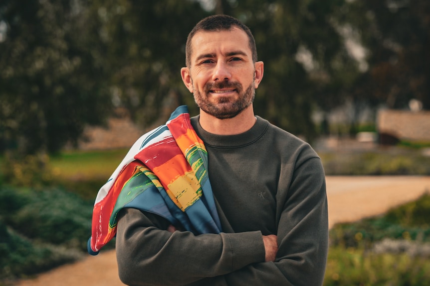 A man with a brown beard and short-cropped haircut poses with a rainbow-coloured garment over his shoulder