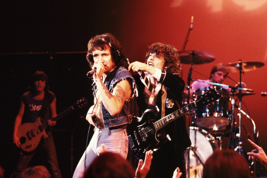 Bon Scott and Angus Young on stage peforming under red lighting setting. Angus puts his necktie around Bon's neck joking fashion