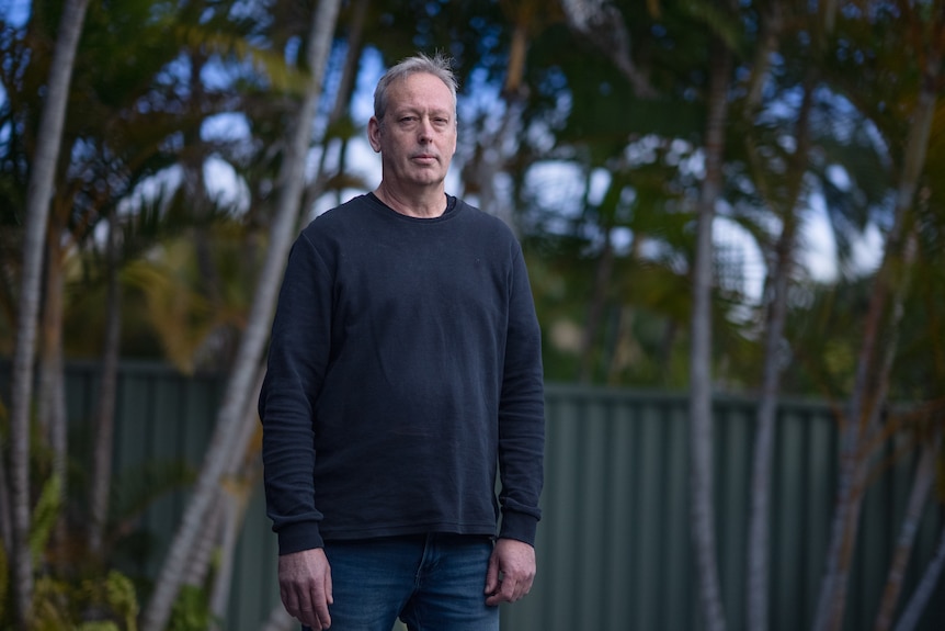 Clay Stevenson stands outside, with palm trees behind him. He looks serious.