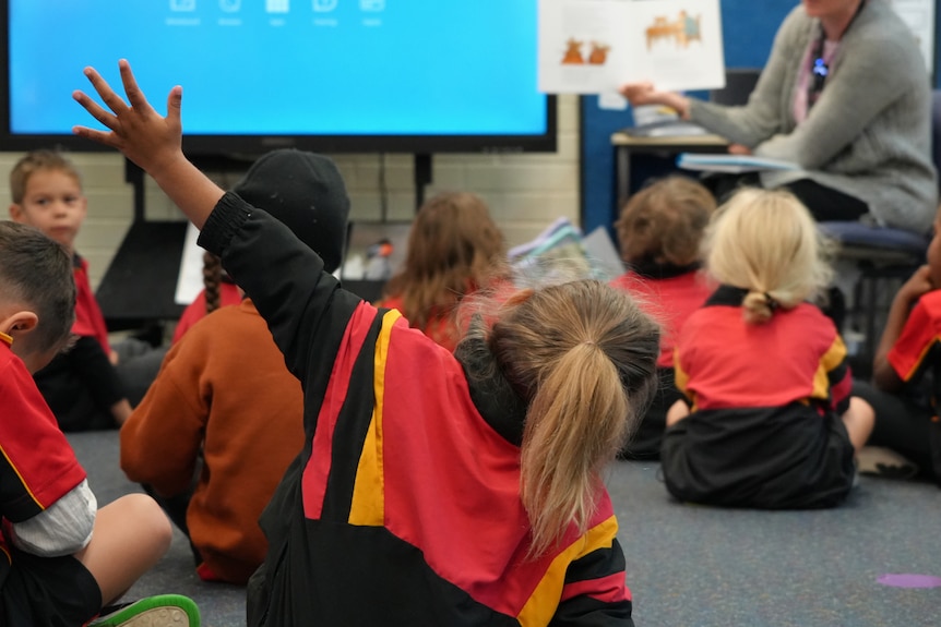 Students in school uniform sitting on the ground in front of a teacher and screen. One has her hand raised in the air.