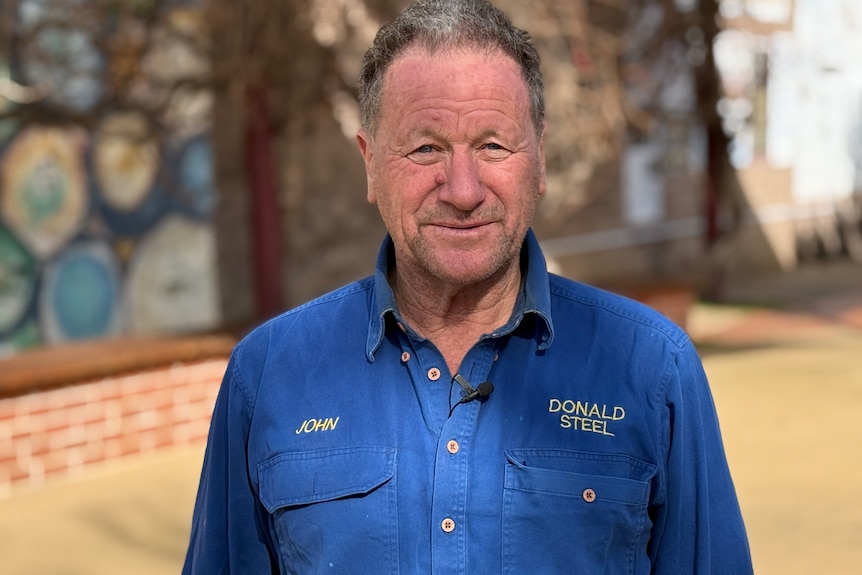 An elderly grey haired man in a navy blue work shirts looks at the camera