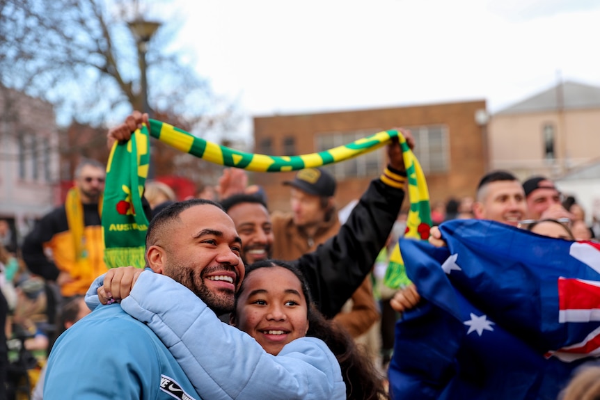 A man and girl hug and smile while a man behind them smiles while holding a green and gold Australia scarf in the air.