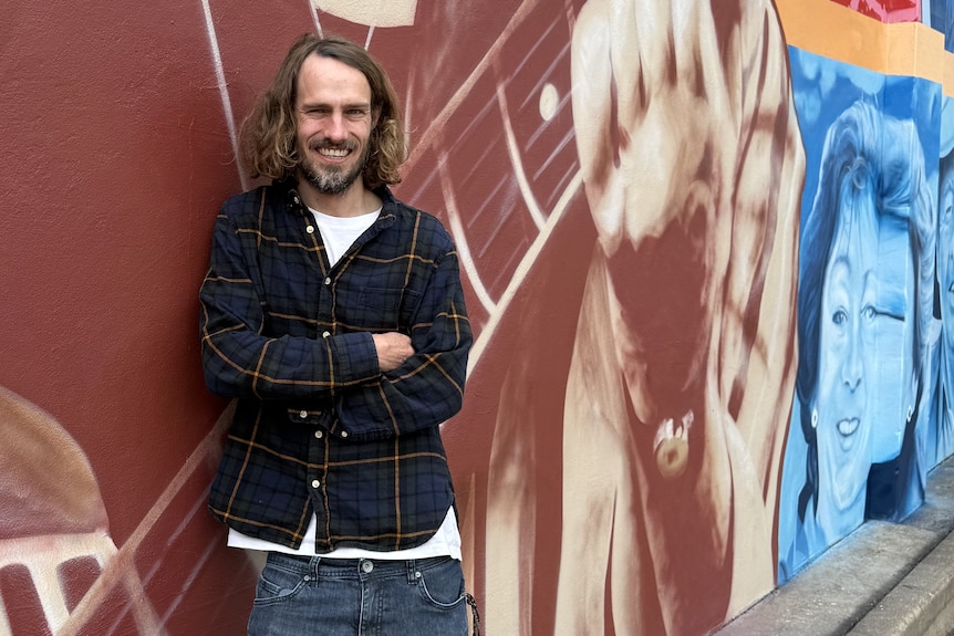 A man with long hair and a beard smiles to the camera leaning against a mural he painted