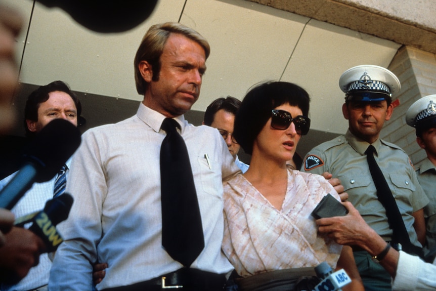 Man and a woman (in a film) interviewed by press outside a police station