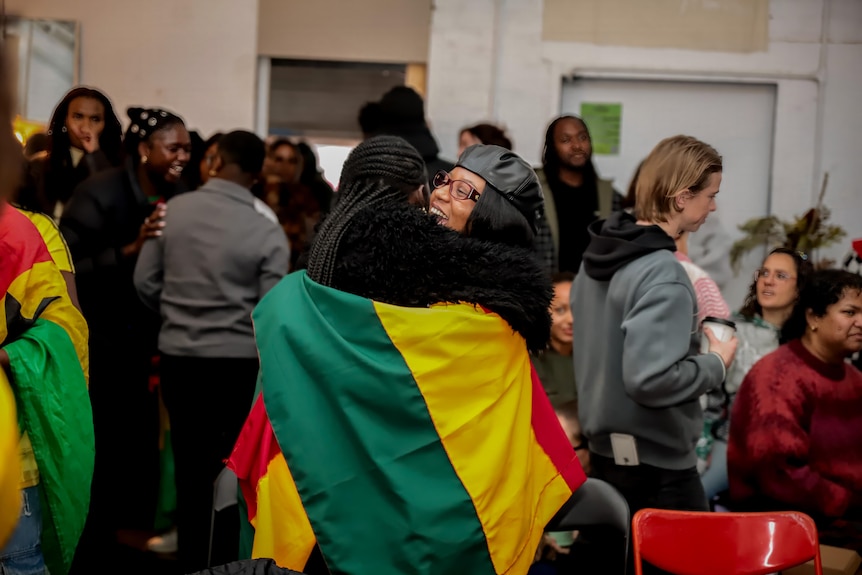 Two people draped in a yellow, red and green flag hug in a room full of people. Others are also wearing red, yellow and green.