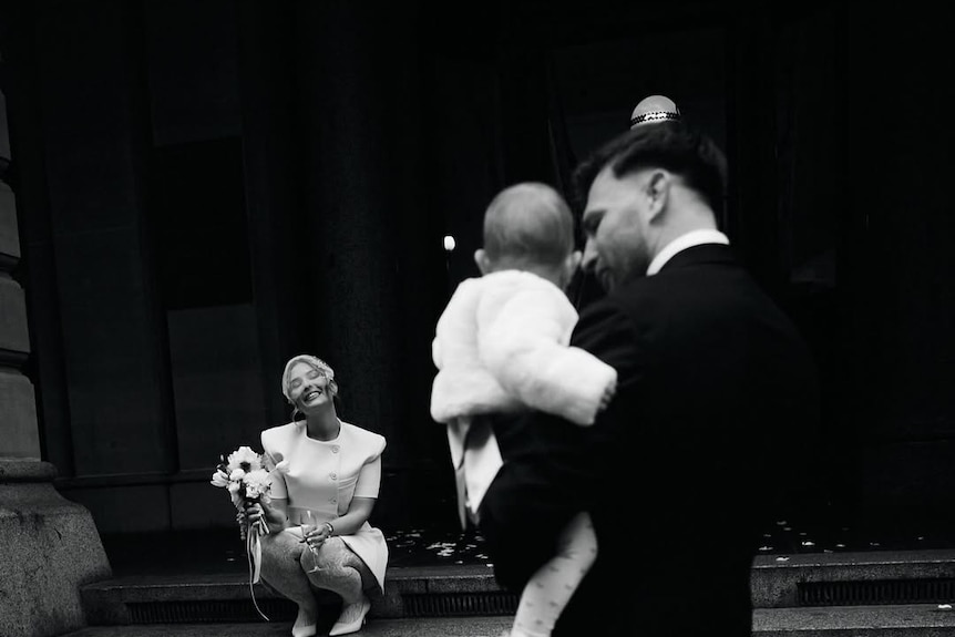 A man holding a baby and looking at a woman in a wedding dress sitting on stairs.