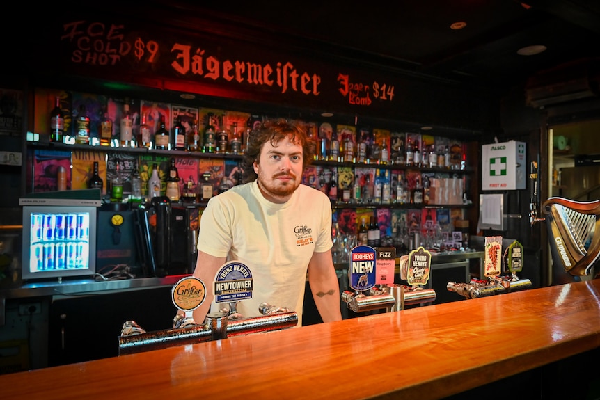 A young man with fair skin and brown hair stands behind a bar.
