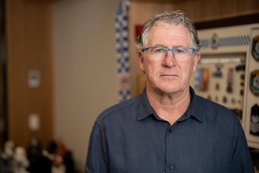 A man with glasses looks at the camera inside an office, a blue and white police logo can be seen in the background.