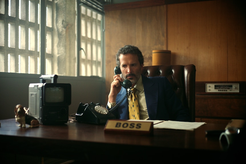 Bret Mckenzie in a vintage suit sits behind a desk on the phone, with a sign on the desk saying BOSS