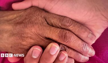A close up of two people holding hands over a pink piece of fabric. The hand on the left is more wrinkly, that the one underneath, on the right.