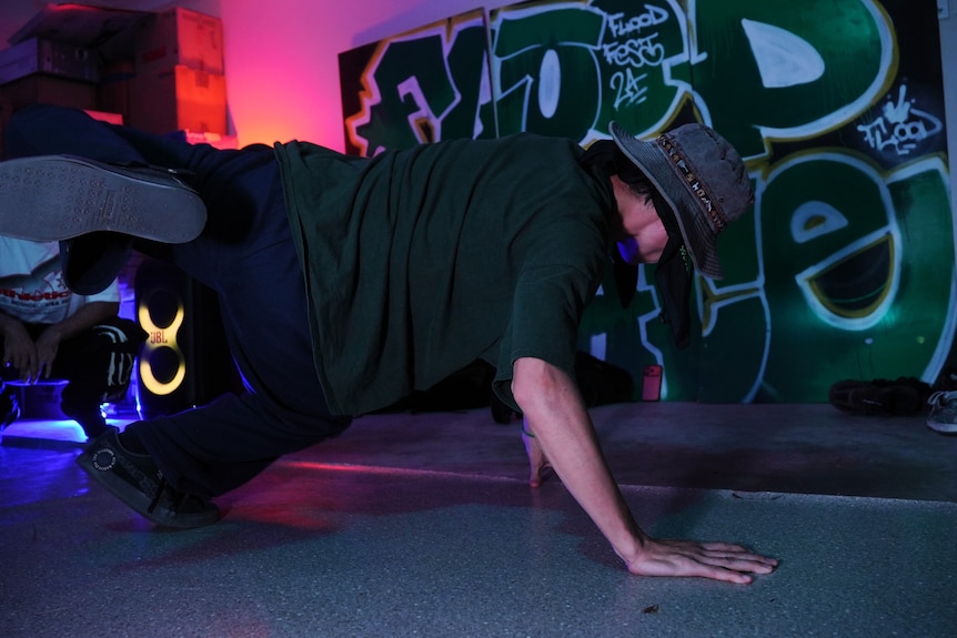 A man wearing a green t-shirt and a bucket hat with three limbs on the floor in the middle of a dance routine.