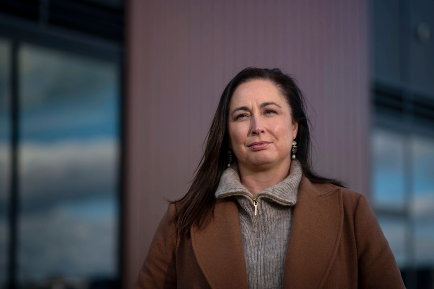 A woman with dark hair and a brown jacket outside metallic office buildings under a blue sky.