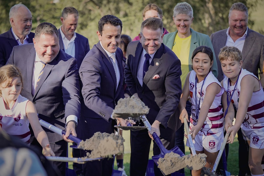 David Crisafulli and Jarrod Bleijie surrounded by children in AFL uniforms lifting shovels with dirt on them.