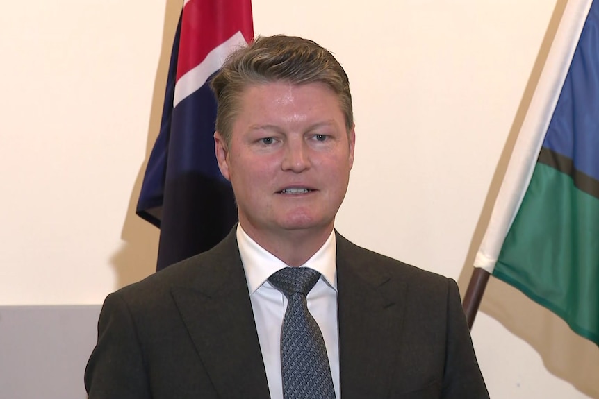 A man with sandy coloured hair in a black jacket, white shirt and blue tie stands in front of two flags.