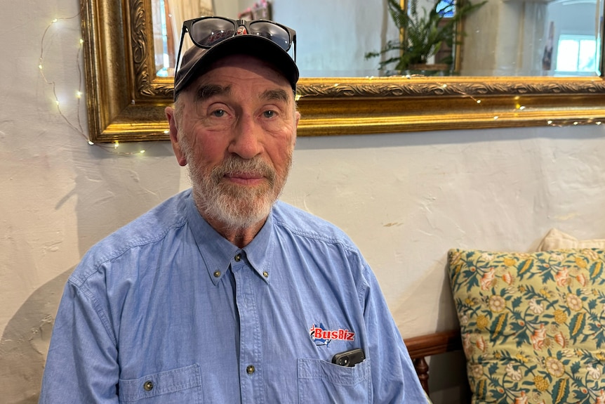 A man with grey hair and a beard wearing a baseball cap inside with a mirror behind him