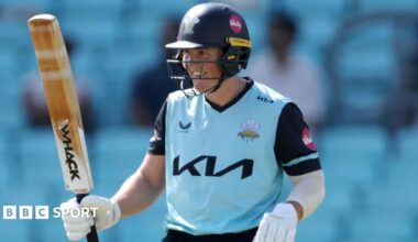 Laura Harris, wearing the Surrey Women's light blue shirt with navy sleeves and navy collar, raises her bat after scoring 50 in 15 balls against Warwickshire Bears