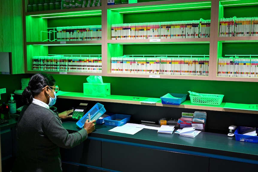 An unnamed pharmacist fills a script near a shelf, coloured by green light, filled with medicines