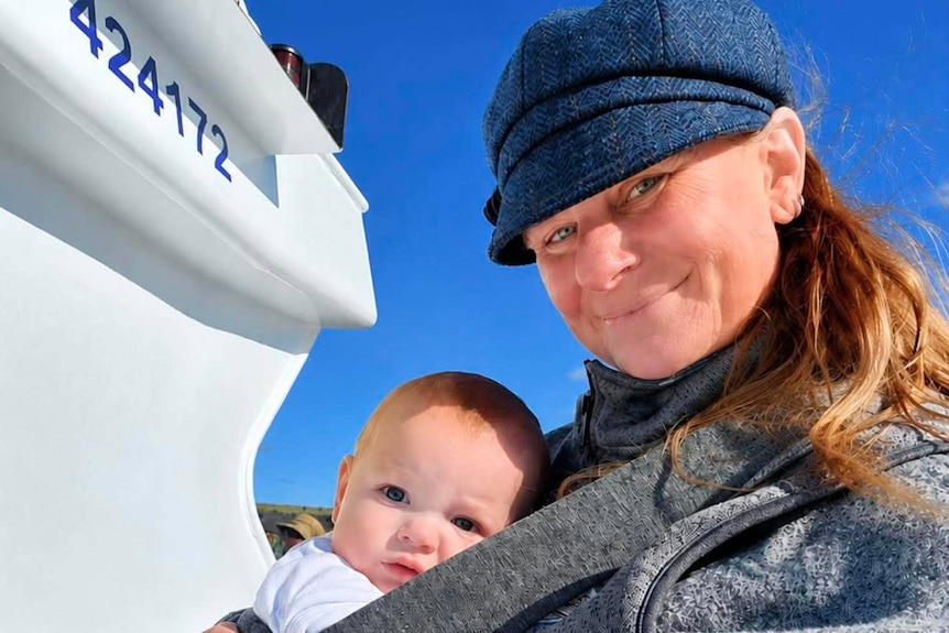 Kim wears a grey jacket and blue hat with a baby strapped to her chest on board a boat.