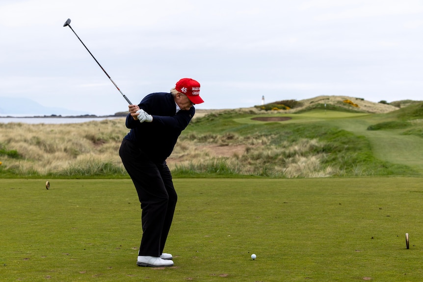 US President Donald Trump takes a swing at a ball on a green golf course.
