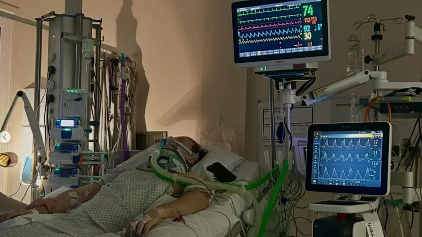 A man lies in a hospital bed attached to several tubes. A series of machines next to the bed shows graphs and squiggly lines