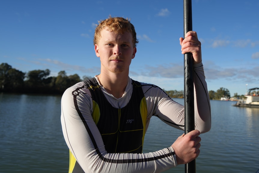 A man in a swim suit holds an oar in front of a river.