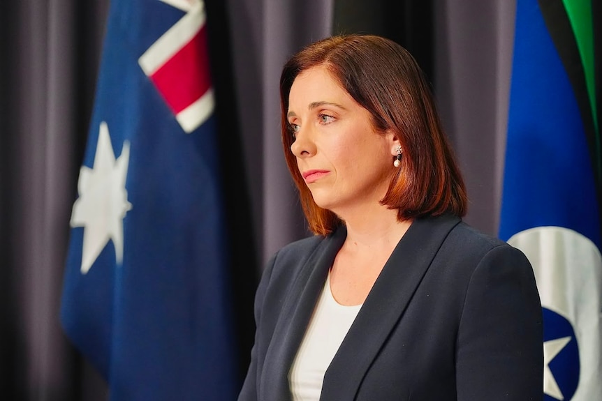 A brown-haired woman wearing a navy blazer, looking solemn.