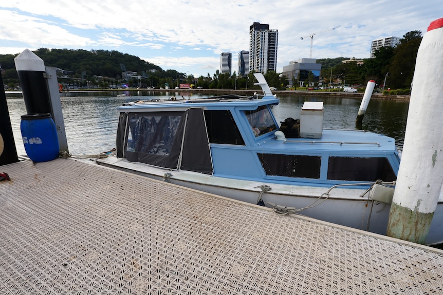 A woman wearing a black hoodie and black pants standing on a wharf next to a blue boat.