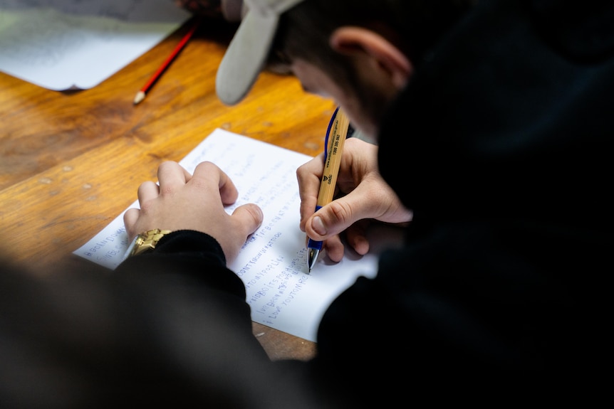 A young man's hands writing on a white piece of paper with a pen, he wears a black hoodie and a white hat.