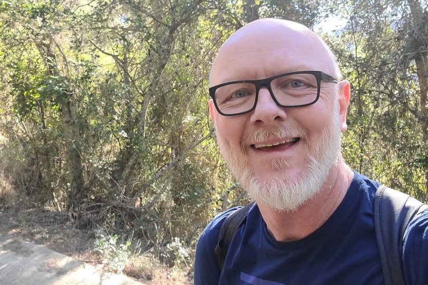 A bald man with glasses and a white beard stands outdoors smiling.