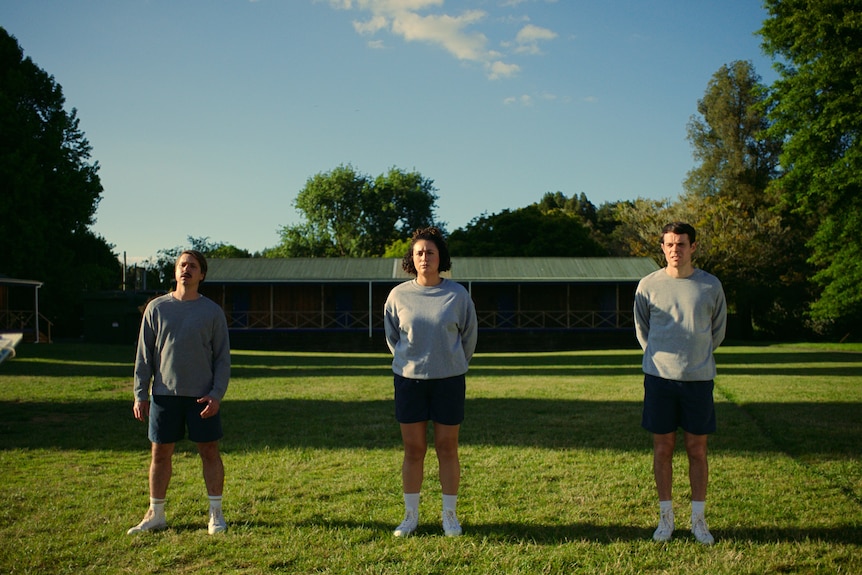 Joe Thomas, Rose Matafeo and Paul Williams stand in a line in athletic shorts and grey jumpers on a green lawn