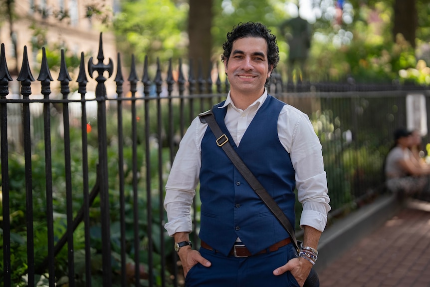 A close up of a man with curly hair wearing a blue vest as he smiles near a garden.