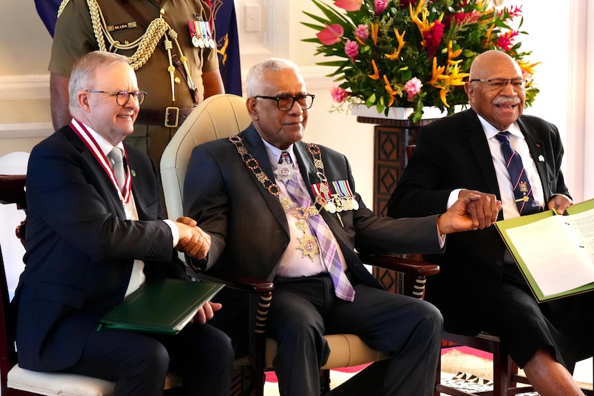 Three seated men hold hands, the two on the sides holding documents in green folders.